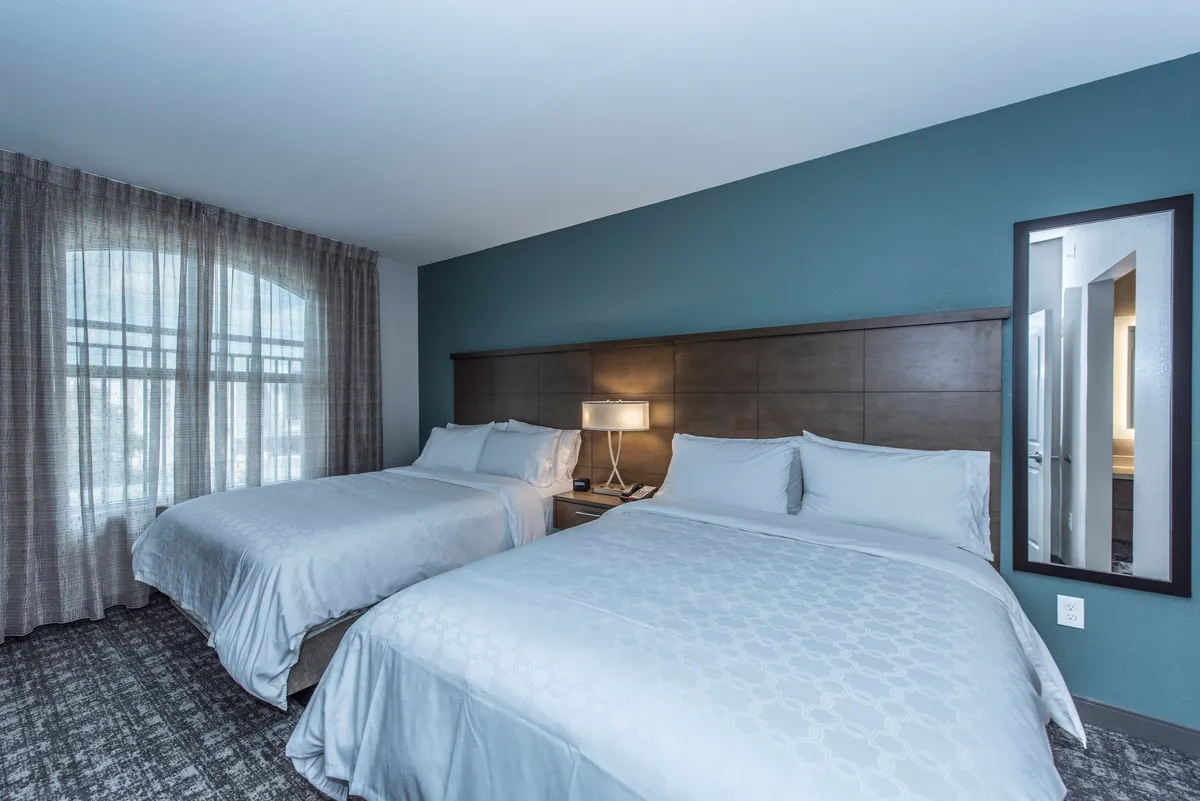 This image shows a cozy bedroom with two queen beds, each with plush bedding and a tasteful, individualized decor style. A nightstand with a lamp sits between the beds, and a large window with blackout curtains ensures privacy and restful sleep. A mirror by the window adds a sense of space and reflects natural light, while the overall setup creates a relaxed, comfortable atmosphere.