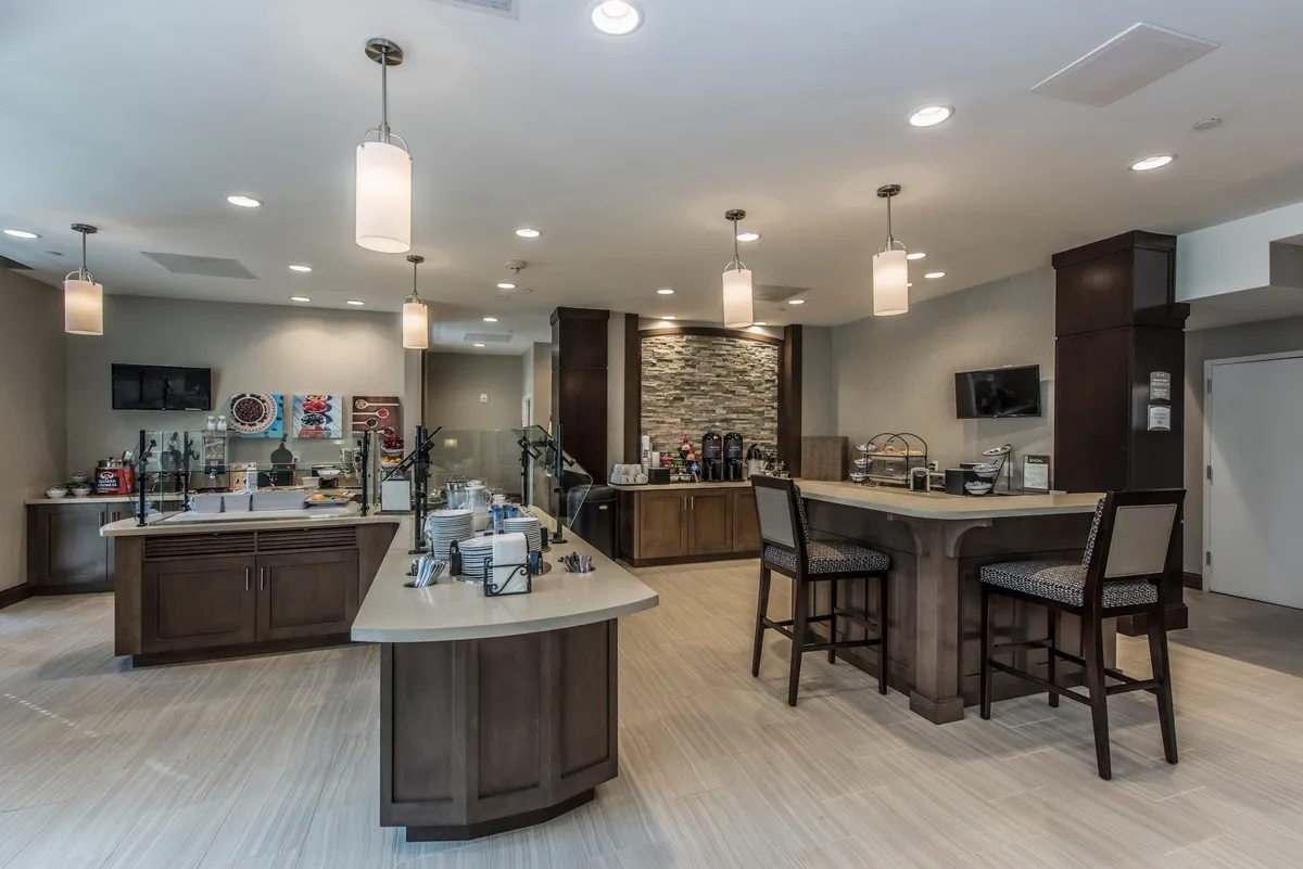 A communal breakfast and beverage area with a central island counter, a dedicated beverage station, and a decorative stone-textured wall. The space is designed for social interaction, casual dining, and morning gatherings, evoking the experience of enjoying a coffee or breakfast with fellow guests.