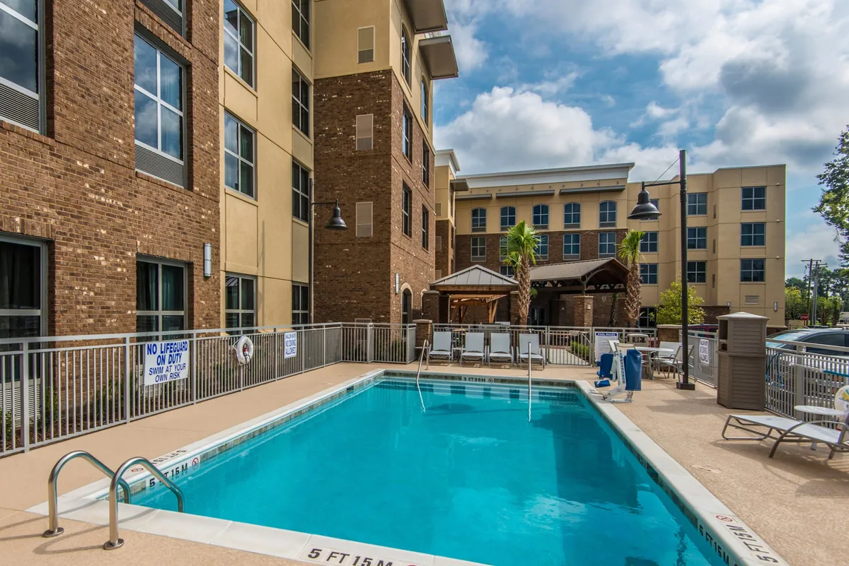 An outdoor swimming pool surrounded by lounge chairs and palm trees, with the building facade visible in the background. The area is set up for swimming, sunbathing, and socializing, creating a relaxed resort-like atmosphere with the possibility of sunset swims and community events.