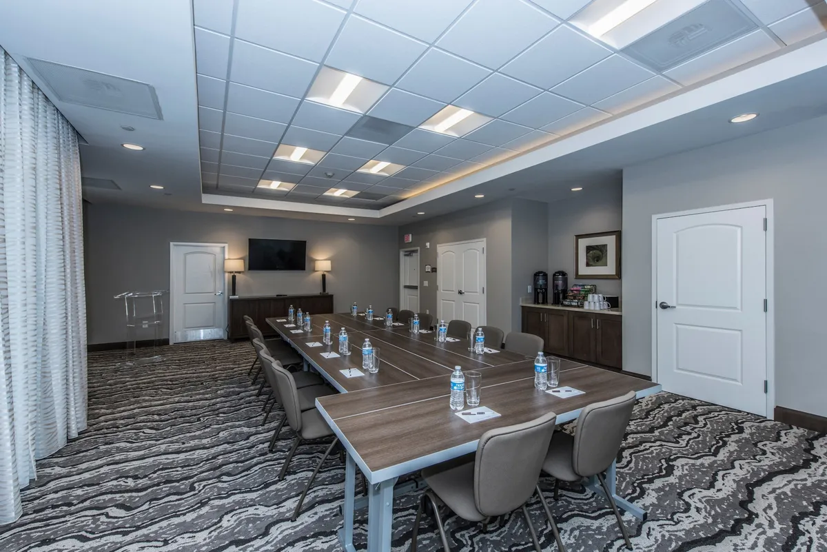 An inviting interior space featuring a long conference table set up for business meetings, with water bottles neatly placed for attendees. The room includes a wall-mounted TV for presentations, decorative artwork on the walls, and doorways that provide a sense of openness. The overall atmosphere is one of focus and collaboration, supporting productivity and teamwork.