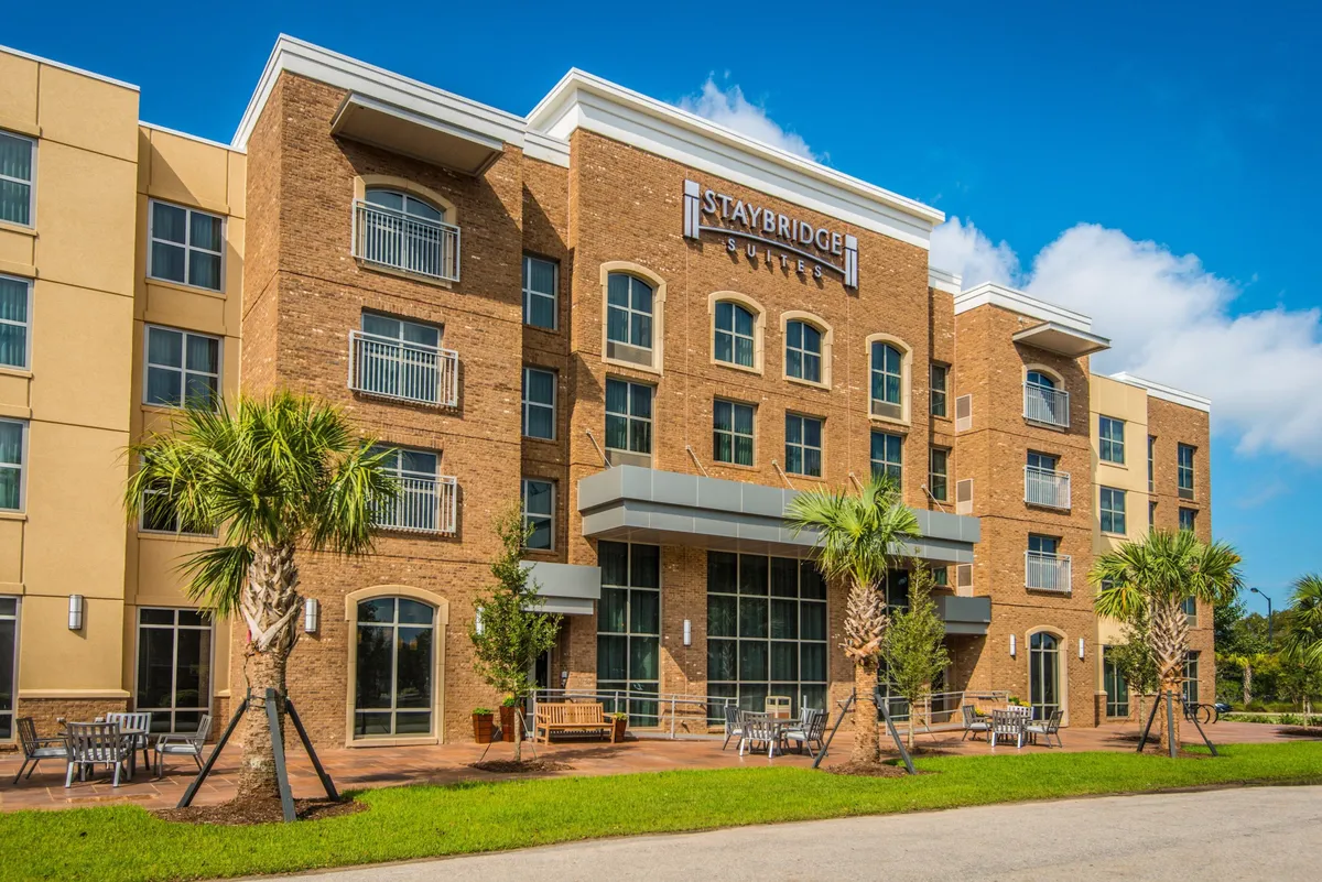 This exterior shot features the Staybridge Suites sign with palm trees and an outdoor seating area. The inviting ambiance is enhanced by sunshine and plenty of space for guests to gather, relax, or enjoy morning coffee outdoors.