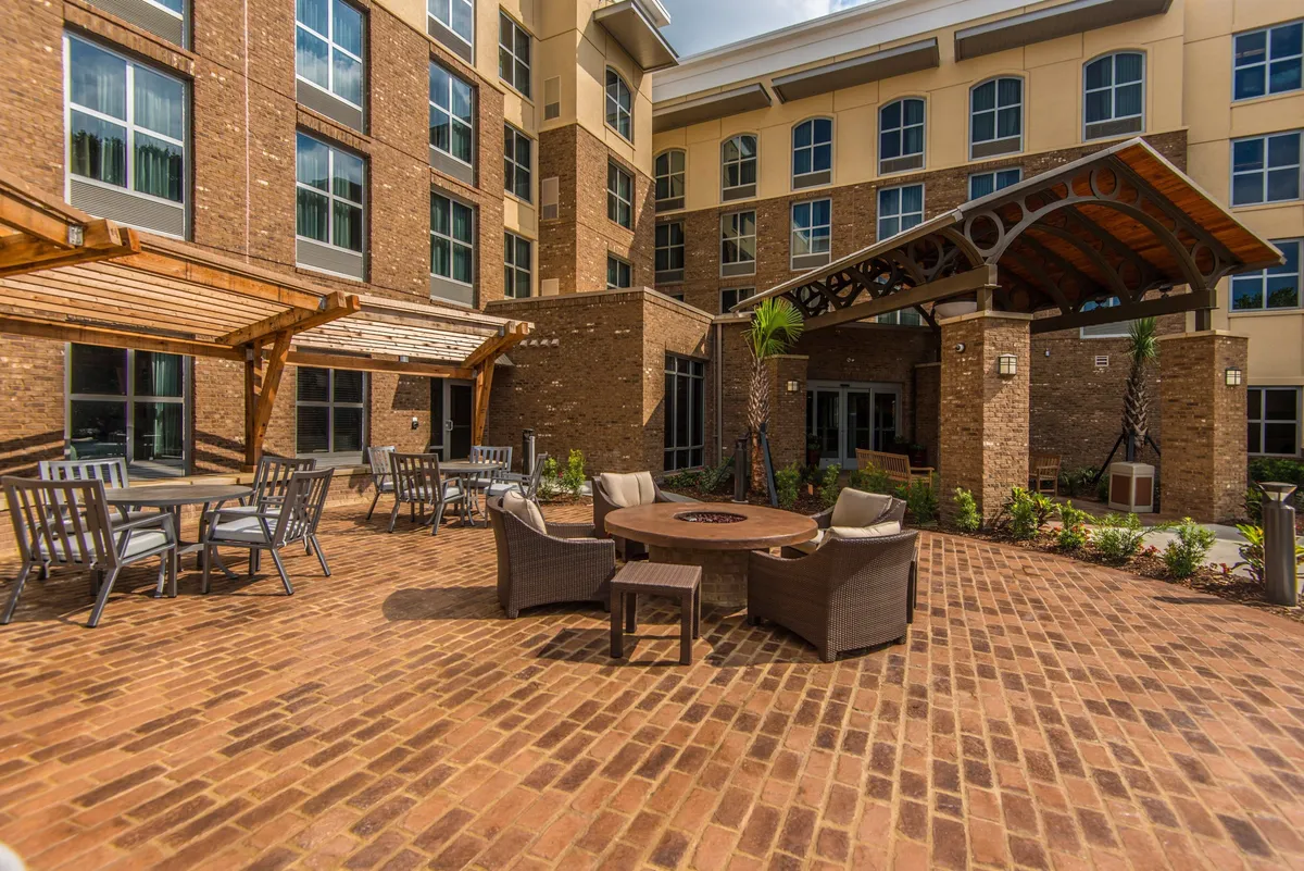 This image highlights a cozy outdoor fire pit area with comfortable seating, landscaped gardens, and the hotel building in the background. The space is ideal for families to gather in the evening, share stories, or enjoy a casual meal outdoors.