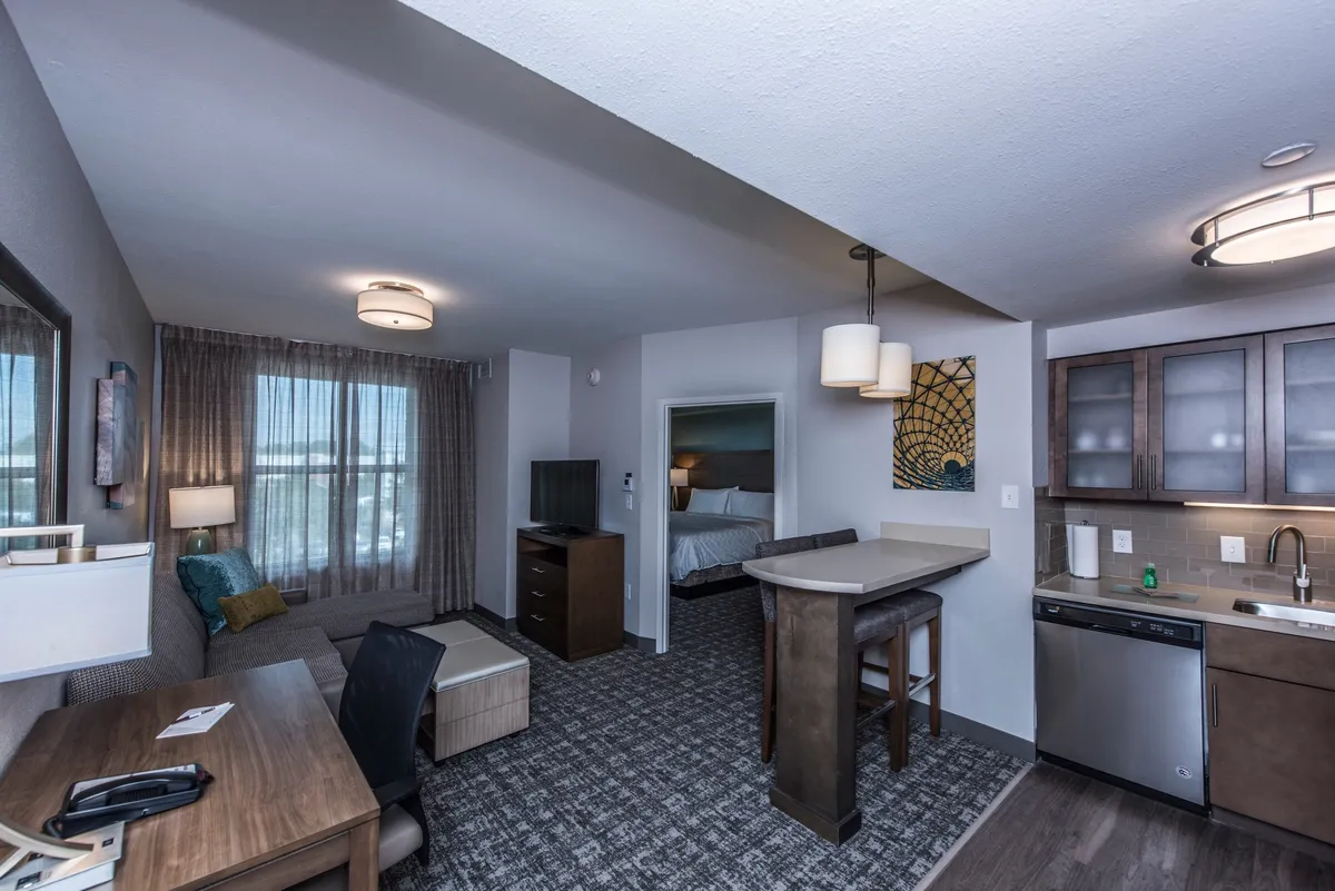 The image showcases a bright, welcoming condo interior featuring a cozy sofa, a modern kitchenette, a practical work desk, and a glimpse into a comfortable bedroom. The layout invites guests to relax, work remotely, prepare meals, or unwind with loved ones in a thoughtfully designed open space.