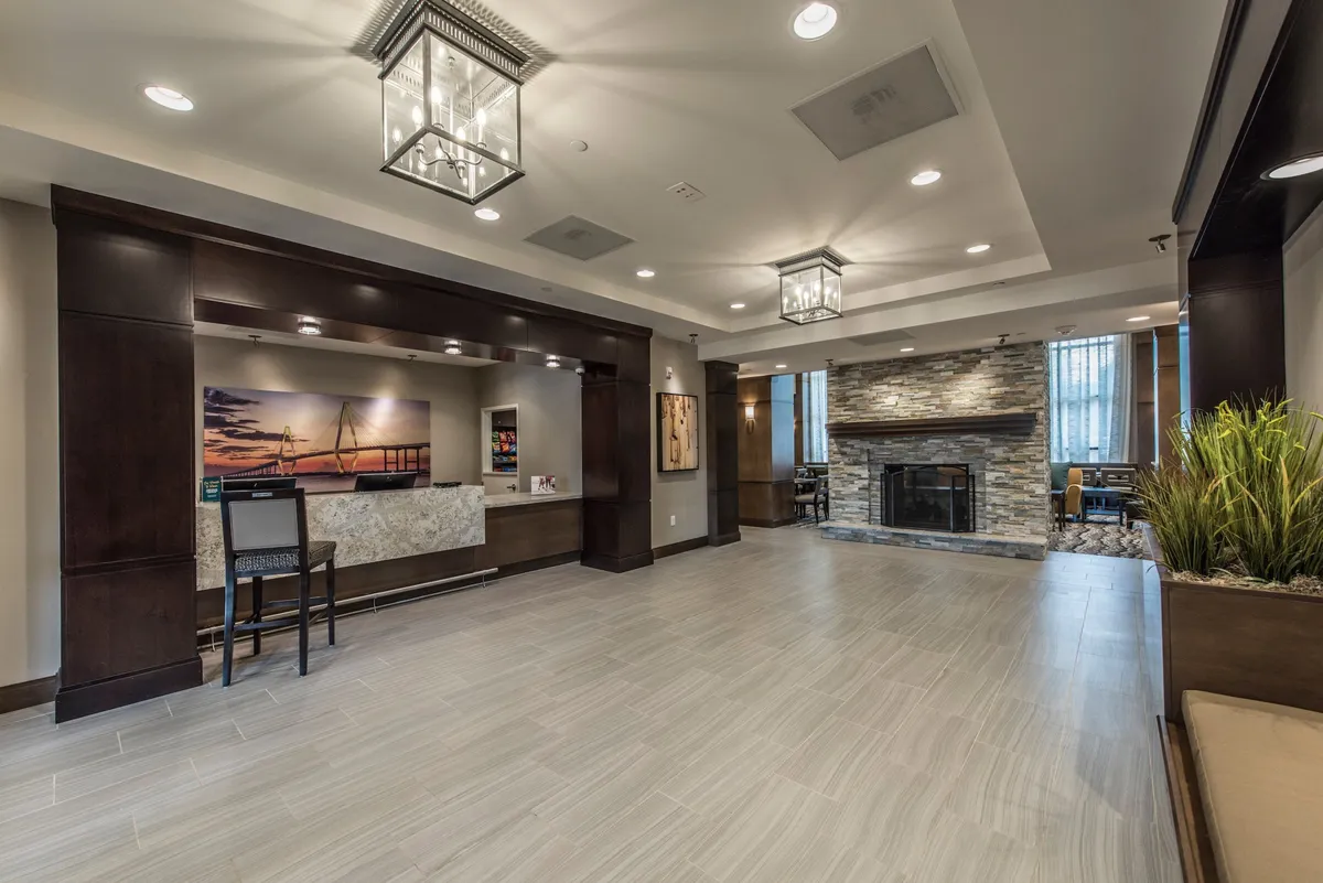 The scene features a stylish living space highlighted by a modern reception desk, a central fireplace, large windows that flood the room with light, decorative plants, and multiple seating areas. Guests are depicted checking in, socializing, and relaxing, underlining the space’s versatility and airiness.