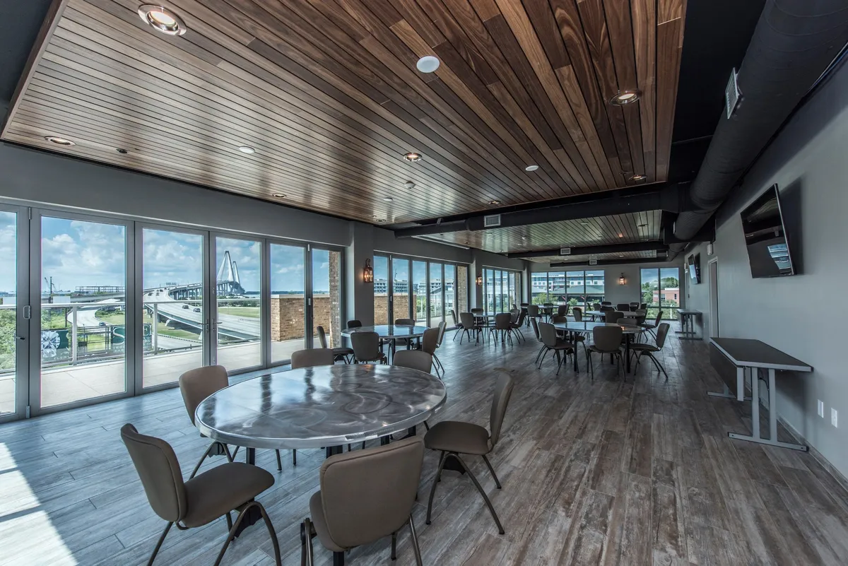 This interior scene features a spacious area with large windows that offer sweeping views to the outside. Round dining tables and a distinctive wooden ceiling set a welcoming tone, while the sunset light creates an inviting and productive atmosphere. The space is arranged to support a variety of activities, including dining, informal meetings, and collaborative work.