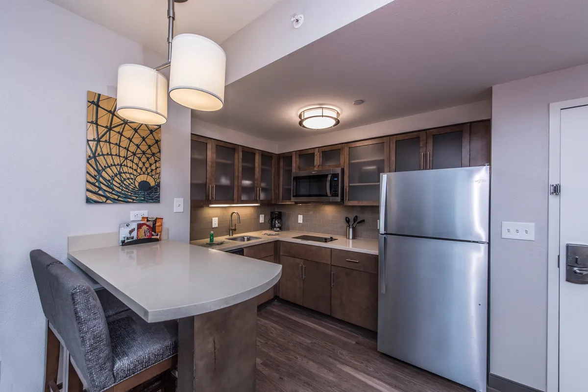 A bright, welcoming kitchen space featuring a central island, a large stainless steel refrigerator, and contemporary lighting fixtures. The setup includes visible countertops and kitchen appliances, suggesting a functional area for cooking, entertaining, or enjoying casual meals. Art on the wall adds a cozy touch, while the arrangement is conducive to both meal preparation and socializing.